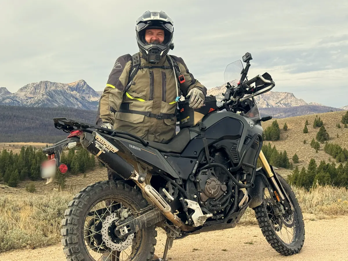 Chad in riding gear and helmet standing behind his adventure bike on a dirt road, jagged mountains behind