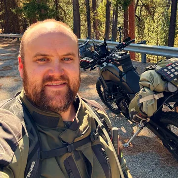 Chad, smiling, in riding gear, his adventure bike loaded with dry bags behind him on a forest road