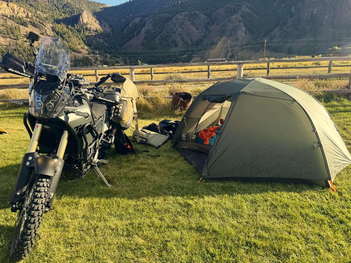 An adventure bike and a small green tent pitched on grass by a rail fence, a canyon wall behind